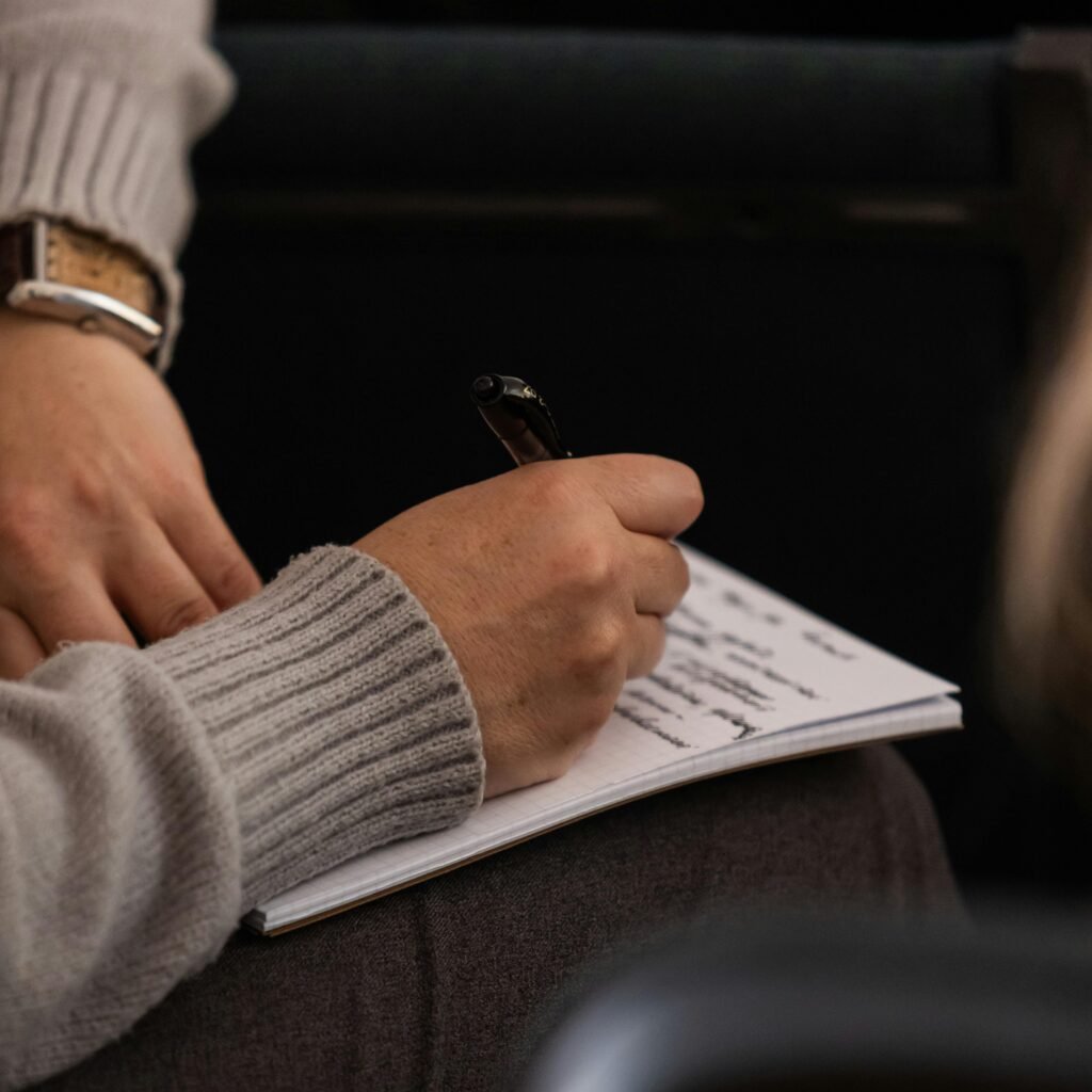 Person in grey sweater taking notes in a notebook with a pen.