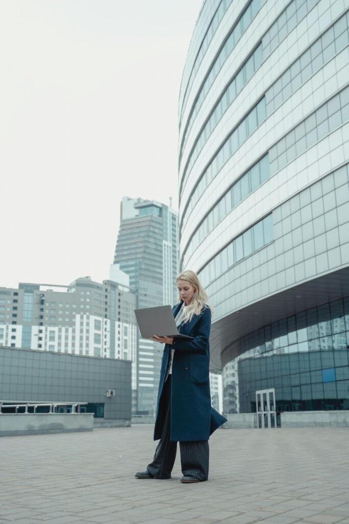 A woman in a business coat working on a laptop outside modern office buildings.